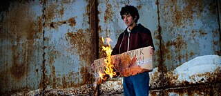 Arshak Makichyan hält ein brennendes Schild aus Pappe auf dem steht „Ihr schafft die Wege zum Klimakollaps“