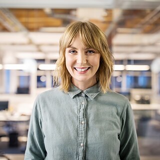 Eine junge Frau in einer Bluse steht in einem Büro und lächelt in die Kamera.
