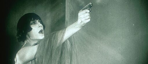 Une jeune femme aux cheveux noirs et courts pointe un pistolet, bras tendu, vers une cible invisible pour le spectateur.