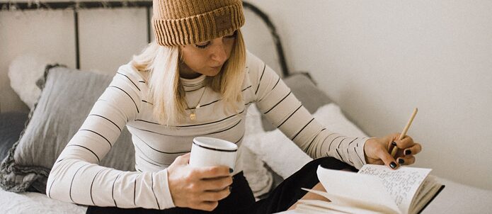 Жінка сидить на ліжку із кружкою в одній руці і блокнотом в іншій