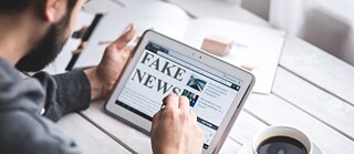 Image of a man holding a tablet, which shows an article with the headline "Fake News"