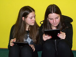 Zwei Mädchen sitzen nebeneinander auf dem Boden und halten Tablets in ihren Händen.