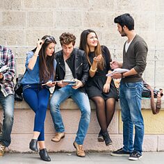Junge Leute sitzen im Freien, haben Bücher und Tablets in der Hand und plaudern. 