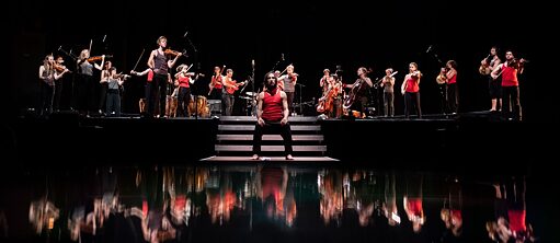 Das STEGREIF.orchester auf einer schwarzen Bühne, die von Wasser umgeben ist. Das Orchester spiegelt sich im Wasser, in der Mitte des Bildes sitzt eine Person auf einem Steg vor der Bühne.