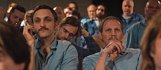 Two men in ligth blue shirts amongs other men in blue shirts looking towards the viewer seemingly watching something
