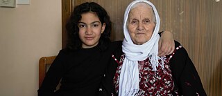  Two women sitting next to each other. On the left 12 year old Thuraya wearing a modern style of clothes. On the right an elderly woman, Um-Reyad, with a traditional embroidered outfit.