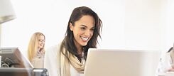 Smiling woman on computer wearing a white blouse and a beige scarf. In the background another woman at the computer out of focus.