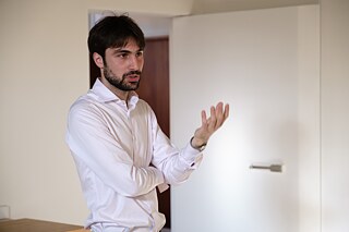 Ένας νεαρός άνδρας με άσπρο πουκάμισο στέκεται μπροστά από μια ανοιχτή άσπρη πόρτα και μιλάει ενώ κάνει χειρονομίες με το αριστερό του χέρι.