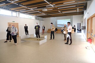 Several people are standing in an exhibition hall. They are all facing towards the camera, as if they are watching something. In the centre of the hall there are two bed frames and at the back a video projection.
