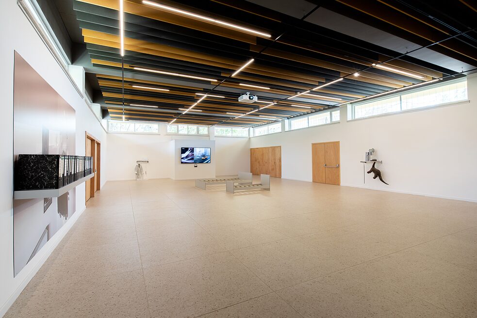 A wide angle view of an exhibition hall. On each of the three walls there is an artwork installation. At the back wall there is a video projection on a television. In the middle of the room are frames of a double bed and a kid's bed.