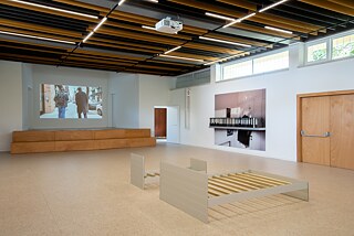 View of two of the walls of an exhibition hall. On the left wall there is a video projection showing the backs of two men. On the right wall there is an art installation with a big print and some boxfiles. There is an open door on the corner where the two walls meet. In the middle of the room are frames of a double bed and a kid's bed.