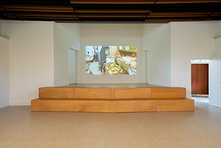 Eine Bühne, vor der sich eine Holzkonstruktion mit zwei großen Stufen befindet. An der Rückwand der Bühne befindet ist eine Videoprojektion, die ein Hasengesicht zeigt, das Gemüse auf einem Markt betrachtet. Auf der rechten Seite des Bildes befindet sich eine offene Tür, die in einen Raum führt, in dem sich eine Lichtinstallation auf dem Boden befindet.