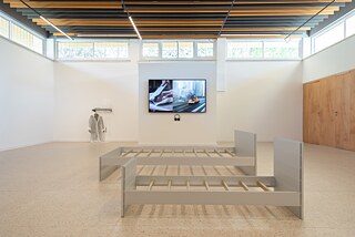 An exhibition hall. In the centre are frames of a double bed and a kid's bed. On the central wall there is a television showing a picture of a car exploding and of two hands cutting food on a cutting board. On the left side of the television there is an art installation with a raincoat. On the right side of the picture there are some wooden doors.