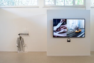 A television, which is hanging on a wall, shows a picture of a car exploding and of two hands cutting food on a cutting board. A set of headphones are hanging below the television. On the left side of the television there is an art installation with a coat and a raincoat hanging from shelves.