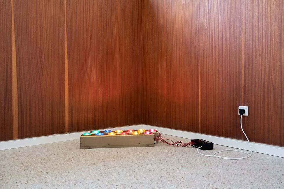 Two rows of colorful lights are placed on the floor of a room, in front of a corner of wood-paneled walls.