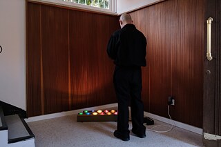 Two rows of colorful lights are placed on the floor of a room, in front of a corner of wood-paneled walls. In front of them stands a man in black clothes with his back turned to the camera.