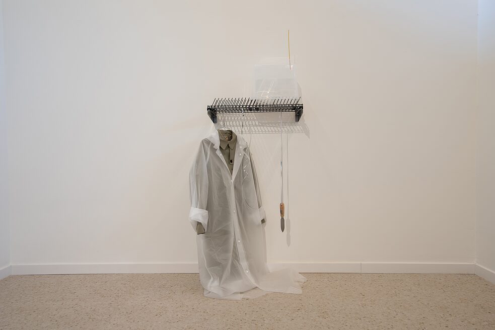 An art installation showing several objects that are connected to each other. A dishwasher rack is fixed on the wall. Several white plastic cans are placed on the right of the rack, with a yellow incense stick on top of them. Below it a scraper is hanging. Next to it, there is a hanger with two coats: a khaki trench coat and a transparent rain coat.