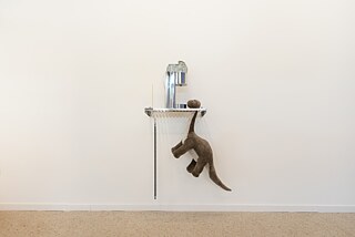 An art installation. A metal rack is attached to a white wall with four objects on it. On the top left there is a yellow incense stick, with a long saw blade hanging underneath it, on the middle of the rack is a kitchen appliance with a silver, reflective surface and on its right a grey and white plush toy in the shape of a dinosaur is hanging off the rack.