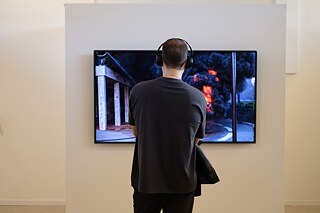 A television placed on a wall shows a video scene where an explosion is happening. A man with headphones is standing in front of the television watching the towards the screen.