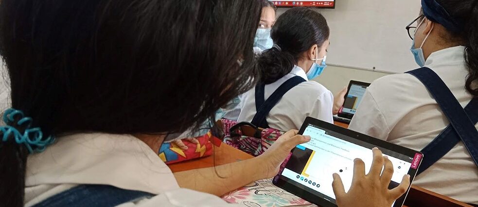 Pupils wearing face masks in a classroom, giving one another feedback on tablets. 