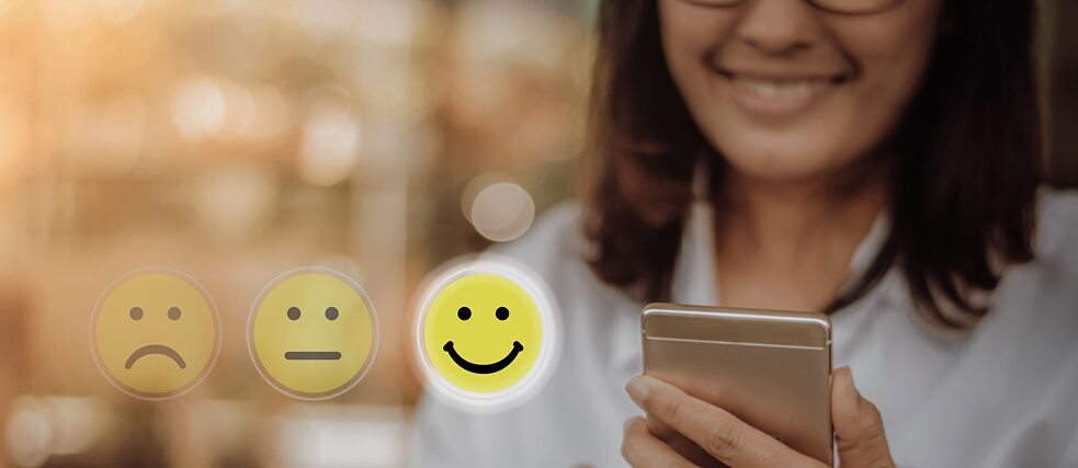 Eine junge Frau sieht auf ihr Smartphone und lächelt. Daneben sind drei Smileys: ein negativer, ein neutraler und ein positiver. Der positive ist hell unterlegt.