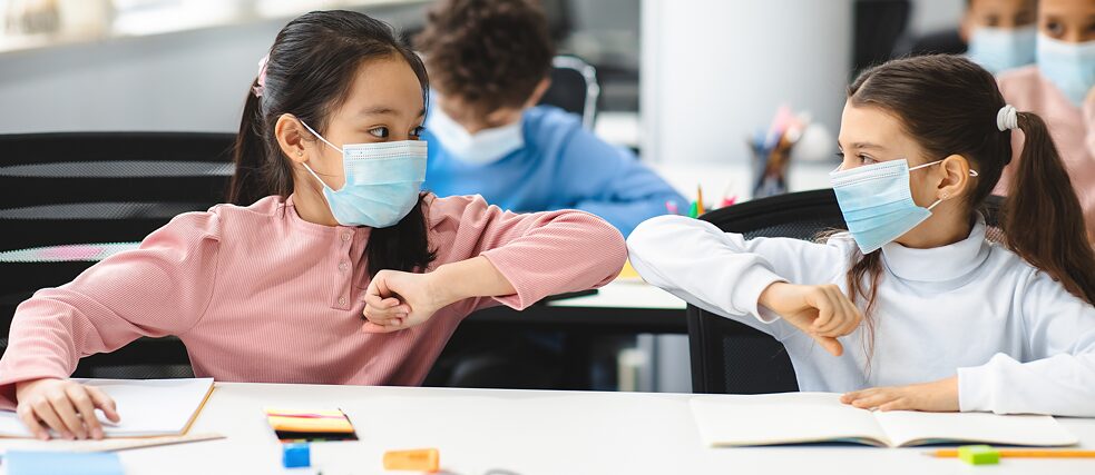Two female students wearing face masks sit at a classroom desk keeping a distance and do an elbow bump.