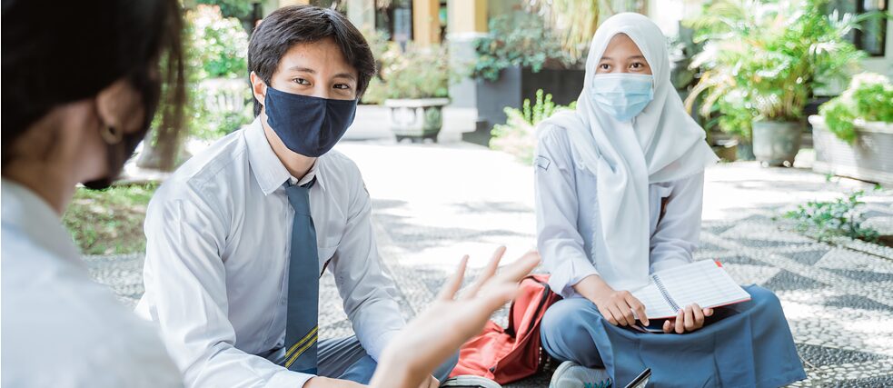 Zwei Maske tragende Schülerinnen und ein Schüler in Schuluniform sitzen auf dem Schulhof auf dem Boden.