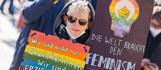 Eine Person mit zwei Protestplakaten mit Botschaften der Unterstützung für Erzieher*innen und Feminismus.