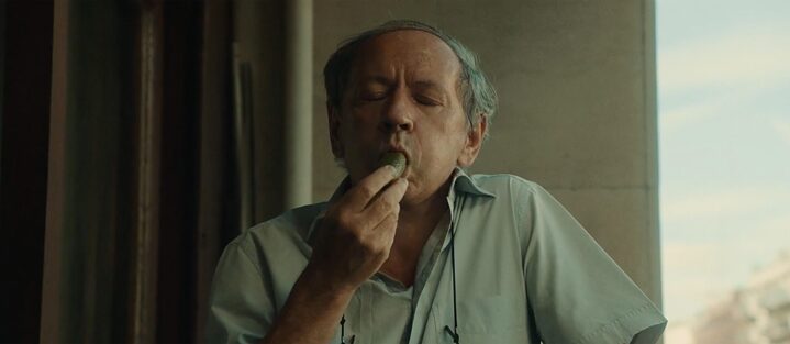 Ein älterer Mann in grau-blauem Hemd beißt genüsslich in eine Gewürzgurke.