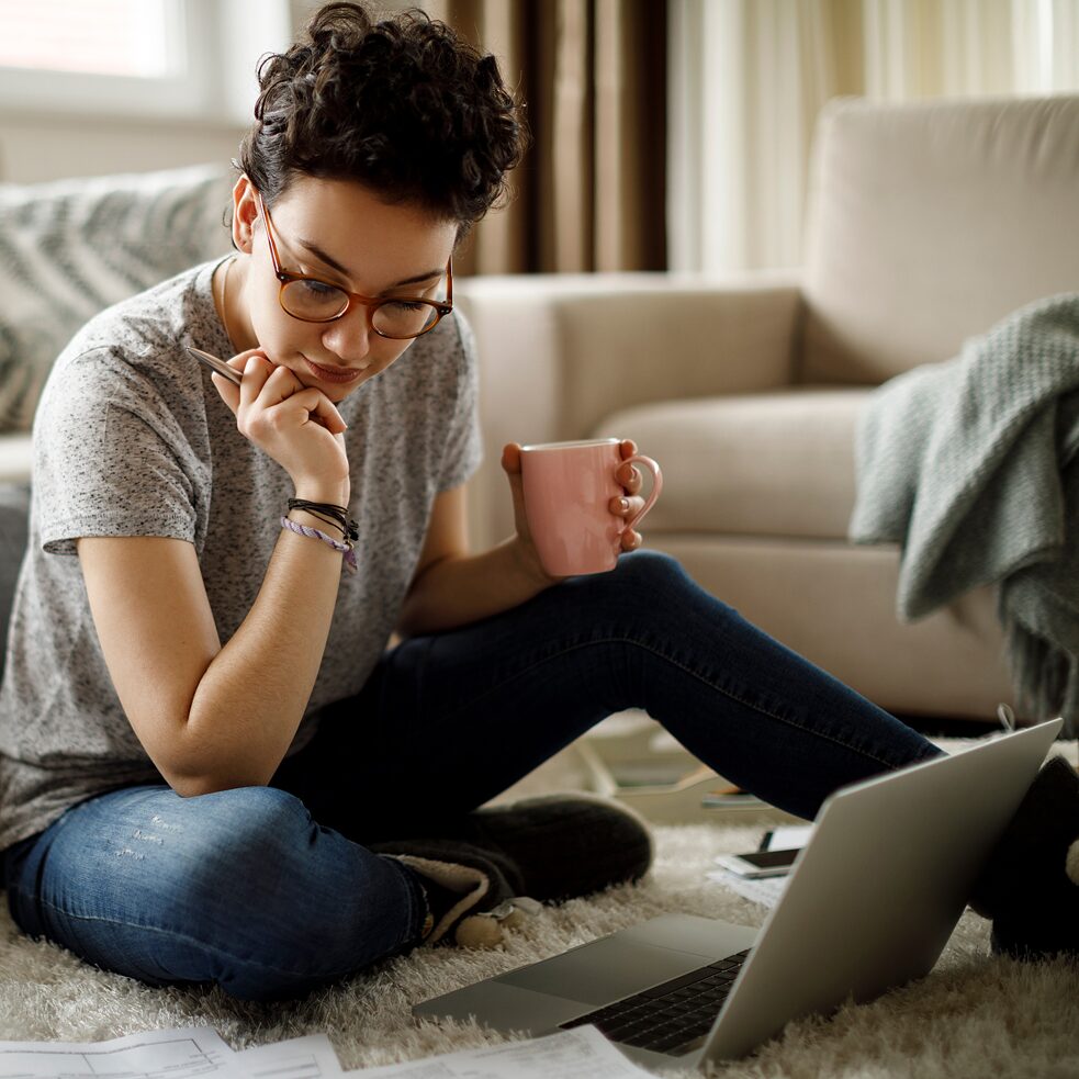 Eine junge Frau sitzt gemütlich zu Hause, am Boden. Vor ihr steht ein Laptop, daneben lieben Unterlagen.