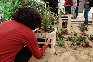 Une jeune femme vue de dos écrit son nom sur un pot de fleurs, fait de palettes.