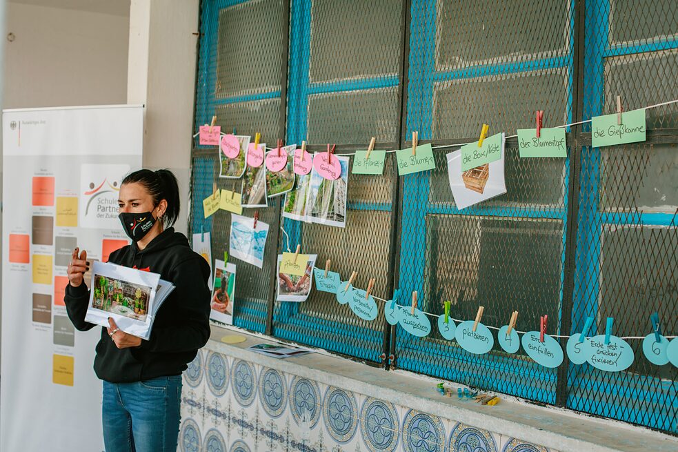 Une femme debout devant une fenêtre sur laquelle est accrochée une corde à linge avec des photos et des mots. Elle tient une photo dans sa main.