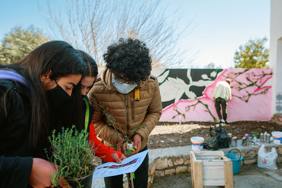 Les élèves tiennent des plantes dans leurs mains et regardent un morceau de papier. En arrière-plan, une personne travaille sur le mur.