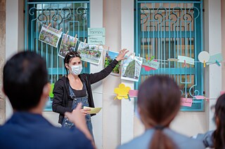 Frau steht vor einem Fenster, an dem eine Wäscheleine mit Fotos und Wörtern hängt. Sie hält ein Foto in der Hand.
