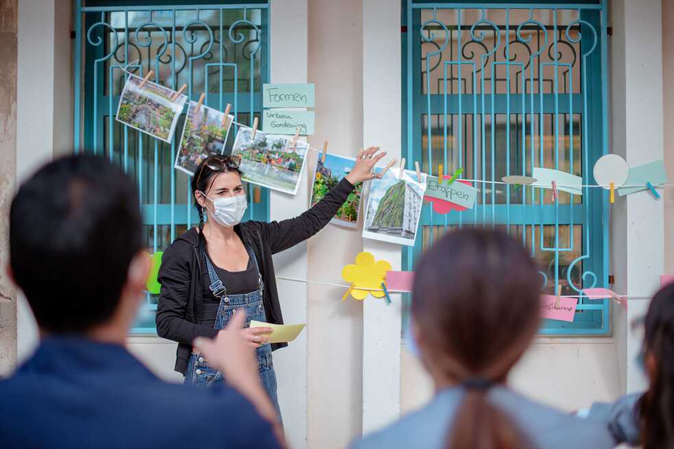 Une femme se tient devant une fenêtre sur laquelle est accrochée une corde à linge avec des photos et des mots. Elle tient une photo dans sa main.