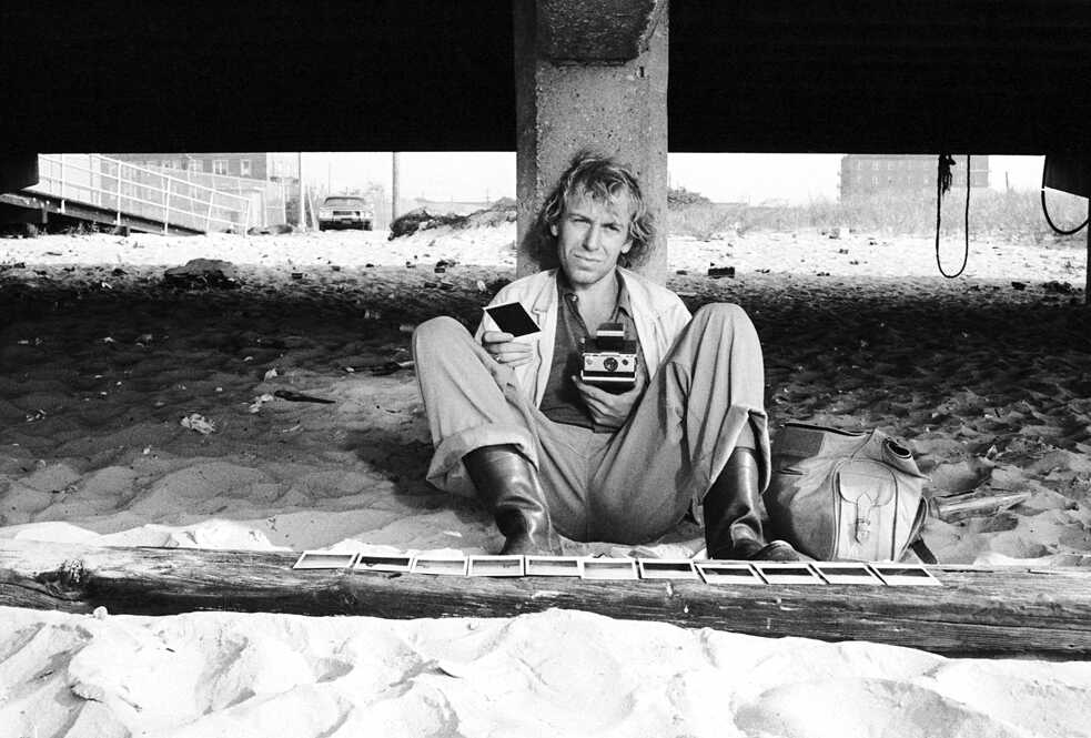 Phillip Winter (Rüdiger Vogler) sitzt in „Alice in den Städten“ am Strand und hält ein Polaroid. 