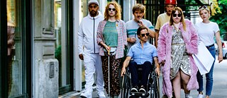 This picture shows seven people walking down the street. On the left is Eddy in a white tracksuit, followed by Magda with sunglasses holding a guide stick. On her right side are Oliver in a wheelchair, and Rainer who has messy hair. Next to them are Michael, wearing a red helmet, and Laura with short blond hair. In front of them walks Franzi, wearing a silver dress and a pink fur coat.