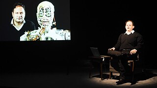 Eine männliche aussehende Figur sitzt auf einem Sessel. Der Hintergrund ist dunkel. Neben ihm ist eine Leinwand, die den menschenförmigen Kopf eines Roboters und einen Mann zeigt.