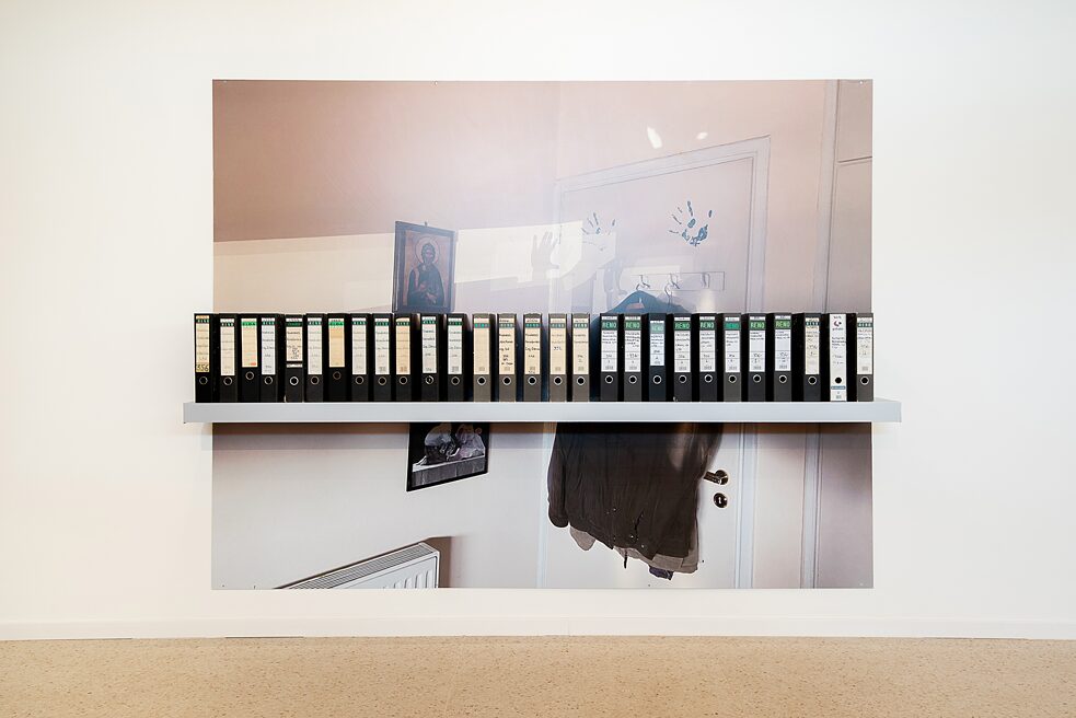 An art installation is attached on a wall. It consists of a big print showing a white door, a tuxedo hanging from the door, a byzantine icon, a framed picture, two handprints, two shadows of open palms and a heating element. In front of the print some black boxfiles are arranged in a row on a shelf.