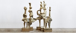 Ein Foto mit weißer Wand im Hintergrund. Im Vordergrund stehen auf einem hölzernen Untergrund fünf bronzefarbene Skulpturen. Die Skulpturen sind versetzt hinter-und nebeneinander platziert und sind alle mit eigenen Bronzesockeln aus dem gleichen Material verbunden. Die Skulpturen ähneln abstrakt menschlichen und tierischen Figuren.