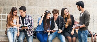 Junge Leute sitzen im Freien, haben Bücher und Tablets in der Hand und plaudern. 