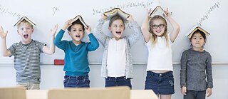 Grundschulkinder stehen vor einer Tafel, sie scherzen und halten Bücher wie ein Dach über dem Kopf.