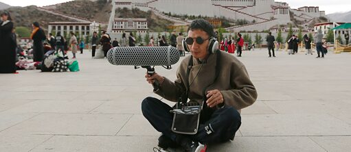 照片中的盲人演说家尼玛旺堆正坐在拉萨布达拉宫广场的空地上，脚边放着他的拐杖。他头戴耳机，右手举着一个巨型麦克风，正在为一个艺术项目录制声音。他的身后站着一群游客。