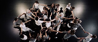 The picture shows a performance by dancers with and without disabilities from an above perspective. The stage floor is black, and six spotlights on the top cast yellowish patches of light on it. The dancers form a complex circle style hand in hand, wearing costumes in black, white, and gray. The work responds to the theatrical scenes in Swiss-born artist Paul Klee’s oeuvre.