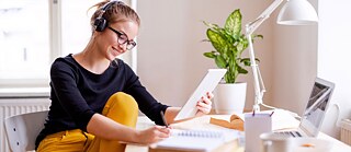 Junge Frau mit Kopfhörern sitzt am Tisch, hält in einer Hand ihren Tablett-PC und schreibt in ihren Notizblock