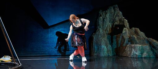  In this staged photo, Julia Häusermann is playing the role of the lead character, Frank. The woman has orange ponytail hair and is wearing a black dress with bright red trousers and white sneakers. The image is in dim blue and black, with the shadow of the heroine and a rockery in the background.