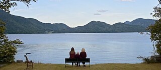 Drei Personen sitzen auf einer Bank an einem See. 