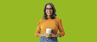 mulher ao centro  da imagem segura copo de café sorri para a camera, atrás vê-se fundo verde sólido, cor da marca Goethe-Institut &copy;    Cursos de alemão online