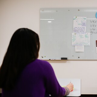 Eine Frau sitzt mit dem Rücken zur Kamera an einem Tisch. Sie hat dunkles Haar und trägt einen violetten Pullover. Sie schreibt mit einem Stift auf ein Blatt Papier, während sie auf das Whiteboard an der Wand vor ihr blickt. Das Whiteboard ist mit deutschen Wörtern aus ihrem Deutschkurs am Goethe-Institut Irland beschriftet.