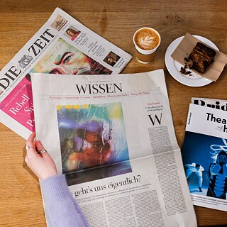 Ein Holztisch ist mit zwei deutschen Zeitungen und einer deutschen Zeitschrift bedeckt. Die linke Hand einer Frau hält eine der Zeitungen; ihr Ärmel ist violett. Neben den Zeitungen, in der oberen rechten Ecke, stehen ein Kaffee und ein Gebäckstück.